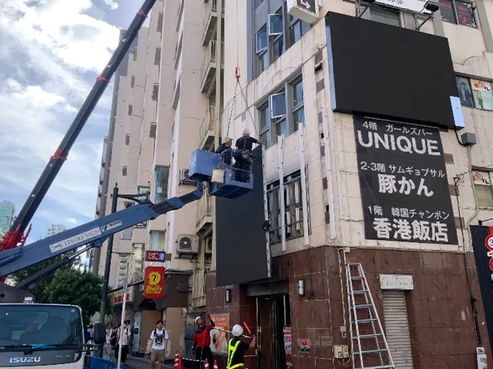 新宿歌舞伎町のビル壁面にクレーンと酵素作業車でLEDビジョンを設置している様子